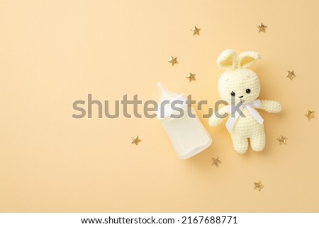 Baby accessories concept. Top view photo of knitted bunny toy milk bottle and gold stars on isolated pastel beige background with copyspace