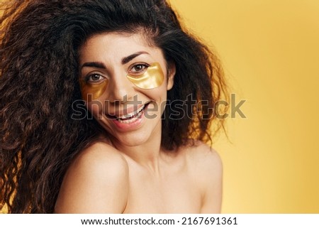 Enjoyed happy pretty Latin female with gold hydrogel patches under eye laughing at camera posing isolated over yellow background. Cosmetic product ad Natural beauty concept Studio portrait