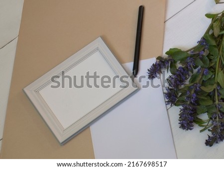 a mockup of an empty photo frame or painting, a sheet of white paper, a black pen, a kraft paper envelope and a bouquet of wildflowers lie on a light wooden table.