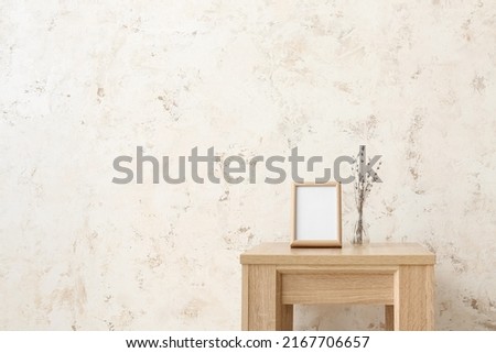 Blank frame and vase with flowers on table near light wall