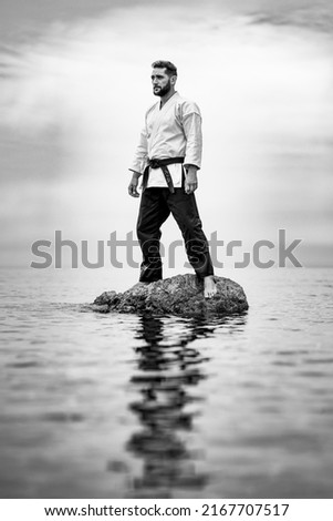 Man standing on a rock in the sea in a karate kimono is a relaxed position with his reflection in the sea water and the Japanese word "Bushido" on his black belt. Royalty-Free Stock Photo #2167707517