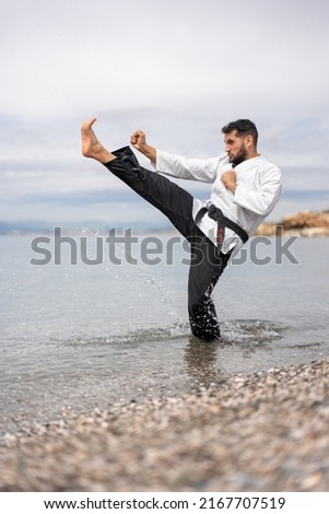 karateka martial arts expert in kimono performing a front kick on the shore of the beach splashing drops of water and on whose belt is written in Japanese "Bushido". Royalty-Free Stock Photo #2167707519