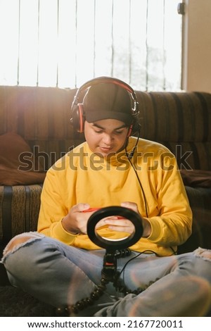 Young man in yellow jacket with headset on his head, looking at cell phone screen with a light in front of him to enhance his online transmission