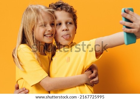  children, brother and sister make selfies on their smartphone showing their tongues to the camera, standing in bright clothes on an orange background. Studio horizontal photography with empty space