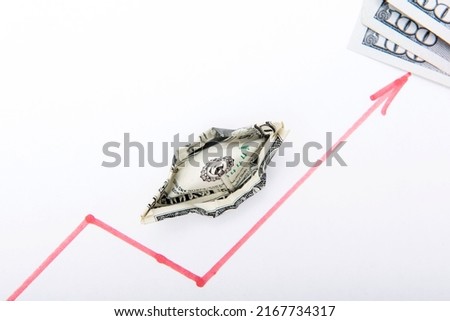 A draw of a chart of rising stock prices on the table together with three dollar banknotes made by red pen are on the white sheet of paper and a small handmade dollar ship. Horizontal studio photo. 