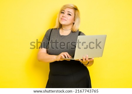 Portrait of a nerdy chubby woman holding a laptop computer while standing against a yellow background