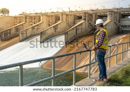 A dam engineering doing his checking routine. He is wearing a white hard hat and yellow transparent vest. He is standing by the rail by the dam. Royalty-Free Stock Photo #2167747783
