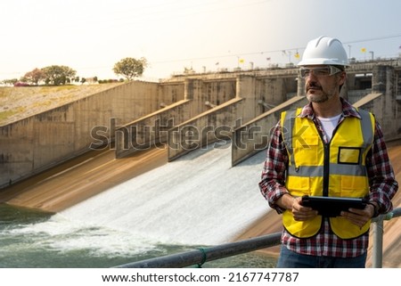 A dam engineering doing his checking routine. He is wearing a white hard hat and yellow transparent vest. He is standing by the rail by the dam. Royalty-Free Stock Photo #2167747787