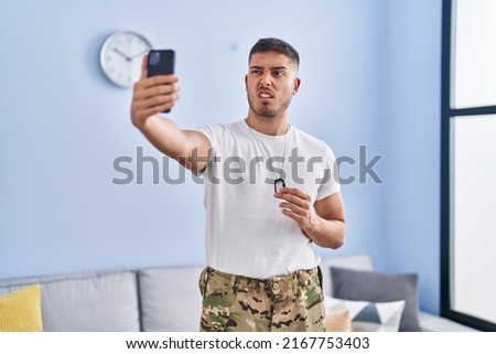 Young hispanic man wearing camouflage army uniform taking selfie at home clueless and confused expression. doubt concept. 