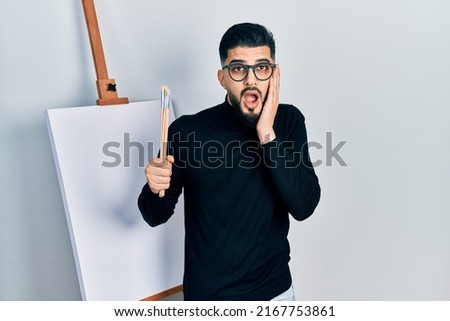 Handsome man with beard holding brushes close to easel stand afraid and shocked, surprise and amazed expression with hands on face 