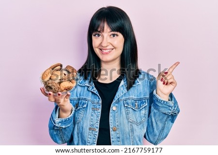 Young hispanic woman holding sweet pastries smiling happy pointing with hand and finger to the side 