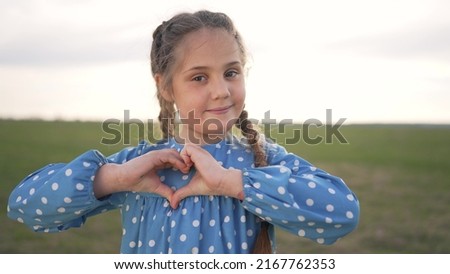 Happy cute girl with kind heart. child makes heart with his hand. Cute smiling little girl in nature makes gesture in shape of heart on green background. concept of child healthy heart, kindness