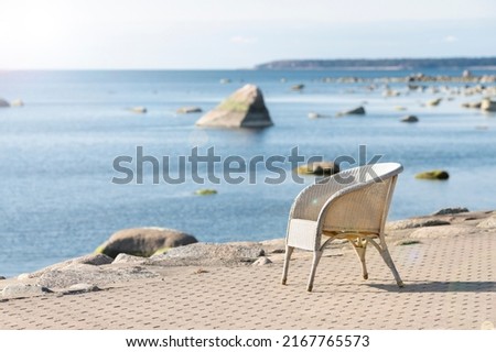 Beach cafe chair with blurred sea background. Summer vacation, travel, holidays concept. Lonely, empty, white wicker seat on shore pavement in sunshine. Seaside restaurant on sunny day. Soft focus Royalty-Free Stock Photo #2167765573
