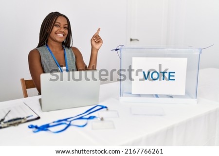 Young african american woman working at political election sitting by ballot with a big smile on face, pointing with hand finger to the side looking at the camera. 