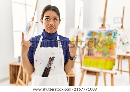 Young brunette woman at art studio pointing up looking sad and upset, indicating direction with fingers, unhappy and depressed. 