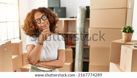 Middle age hispanic woman wearing casual clothes standing around cardboard boxes at new home serious face thinking about question with hand on chin, thoughtful about confusing idea 