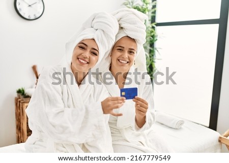 Woman couple wearing bathrobe holding credit card sitting on massage board at beauty center
