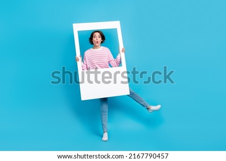 Full size photo of impressed pretty girl fooling around with big white snapshot frame isolated on blue color background