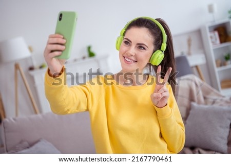 Photo of influencer pretty lady hold telephone make selfie show v-sign wear earphones yellow shirt comfortable home indoors