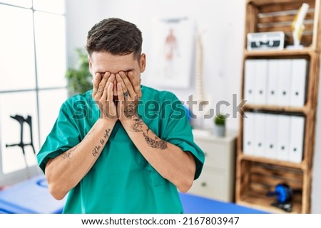 Young physiotherapist man working at pain recovery clinic with sad expression covering face with hands while crying. depression concept. 