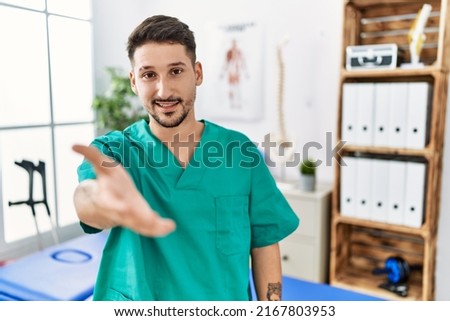 Young physiotherapist man working at pain recovery clinic smiling cheerful offering palm hand giving assistance and acceptance. 