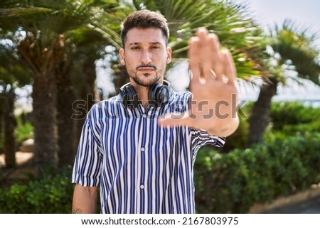 Young handsome man listening to music using headphones outdoors doing stop sing with palm of the hand. warning expression with negative and serious gesture on the face. 