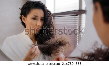Upset sad young woman distressed by damaged hair frustrated about split ends girl worried about loss brittle dry hair hormone problem vitamin deficiency reflecting in mirror haircare treatment concept Royalty-Free Stock Photo #2167804383