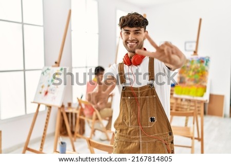 Young hispanic man at art studio smiling looking to the camera showing fingers doing victory sign. number two. 