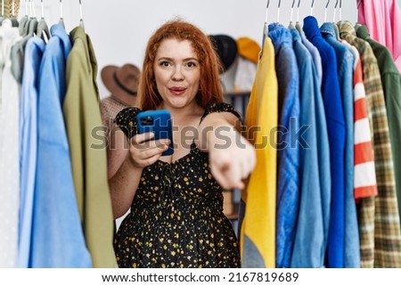 Young redhead woman searching clothes on clothing rack using smartphone pointing with finger to the camera and to you, confident gesture looking serious 
