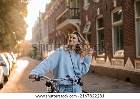 Pretty young woman on bicycle in the city street, city transportation. Outdoor fashion portrait of elegant blonde curly lady riding her hipster retro bike in stylish blue sweater. Girl fix hir.