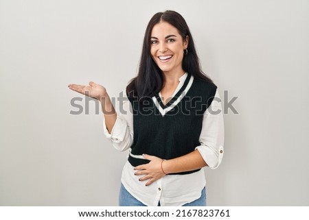 Young hispanic woman standing over isolated background smiling cheerful presenting and pointing with palm of hand looking at the camera. 