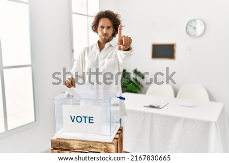 Young hispanic man voting putting envelop in ballot box pointing with finger up and angry expression, showing no gesture 
