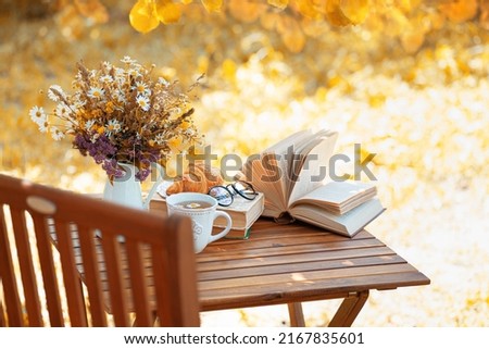 Bouquet of flowers, croissant, cup of tea or coffee, books on table in autumn garden. Rest in garden, reading books, breakfast, vacations in nature concept. Autumn time in garden on backyard Royalty-Free Stock Photo #2167835601