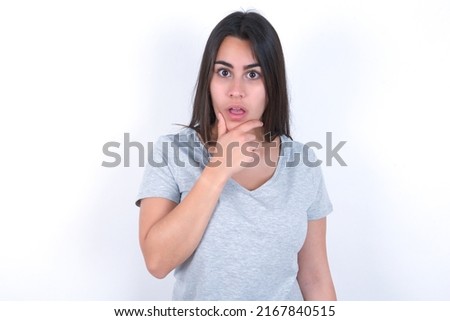young caucasian woman wearing grey t-shirt over white background Looking fascinated with disbelief, surprise and amazed expression with hands on chin