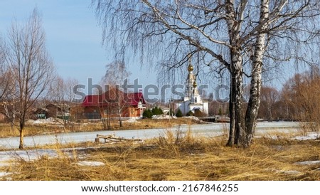 White Church in name of Holy Prince Alexander Nevsky, Baltym village, Ural, Sverdlovsk region, Russia. Village administration, birch tree without leaves, yellow grass and river or pond covered ice.