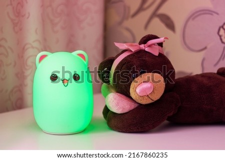Baby cute green bear-shaped night lamp with eyes and ears near a toy teddy bear with a bow on its head and a heart in its hands on the bedside table in the pink baby girl room glowing in the dark