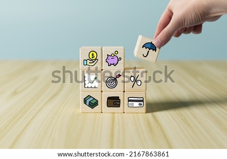 The hand adds a wooden cube with an umbrella icon next to the icons of saving, money, wallet, piggy bank. Concept of taking care of your investments, money. Safeguard.