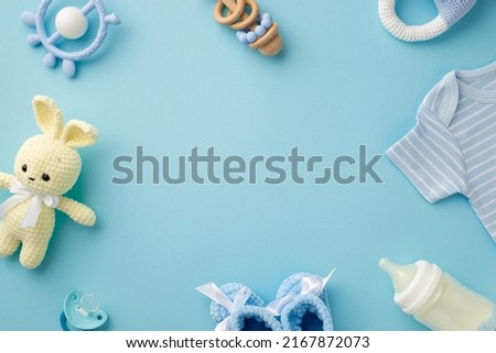 Baby accessories concept. Top view photo of infant clothes blue shirt knitted booties bunny toy teether rattle bottle and pacifier on isolated pastel blue background with empty space in the middle