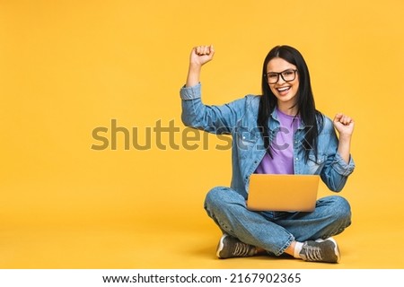 Happy winner! Business concept. Portrait of happy young woman in casual sitting on floor in lotus pose and holding laptop isolated over yellow background.