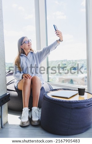 Freelancer woman in classic glasses talking on the phone and smiling while sitting in a modern coworking space, carefree millennial woman in glasses enjoying leisure time for communication