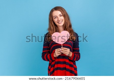 Kind friendly woman wearing striped casual style sweater, holding pink heart on stick, looking at camera with smile, expressing love. Indoor studio shot isolated on blue background.
