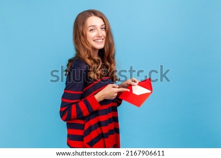 I got love letter on Valentine's day. Woman in striped casual style sweater, pointing letter in red envelope, holding greeting card and smiling joyfully.Indoor studio shot isolated on blue background. Royalty-Free Stock Photo #2167906611