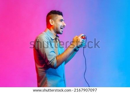 Side view of man in shirt holding in hands red gamepad joystick, grimacing playing video games with happy facial expression. Indoor studio shot isolated on colorful neon light background.