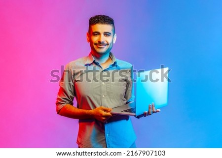 Portrait of happy positive man in shirt holding notebook in hands, looking at camera with positive facial expression. Indoor studio shot isolated on colorful neon light background.