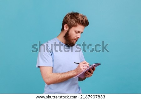 Portrait of bearded man making notes in paper notebook, writing business idea, future plans, checking appointment in schedule diary. Indoor studio shot isolated on blue background. Royalty-Free Stock Photo #2167907833