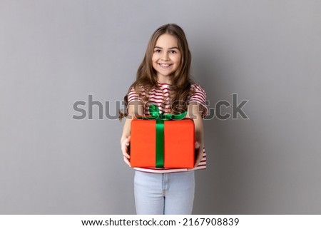 Portrait of smiling little girl wearing striped T-shirt giving present and congratulating on birthday, mother's day, offering gift box for charity. Indoor studio shot isolated on gray background.