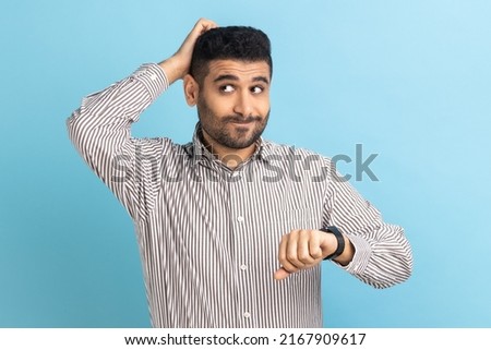 Confused businessman standing with puzzled facial expression, worried about deadline, looking away, keeps hand on head, wearing striped shirt. Indoor studio shot isolated on blue background. Royalty-Free Stock Photo #2167909617
