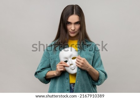 Portrait of serious bossy woman holding white mask in her hand, changing personality, hiding mask in the bosom, wearing casual style jacket. Indoor studio shot isolated on gray background. Royalty-Free Stock Photo #2167910389