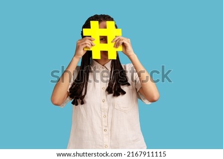 Funny woman blogger or smm manager with dreadlocks hiding face behind yellow hashtag sign, teaching to tag topics on websites, giving recommendations. Indoor studio shot isolated on blue background.