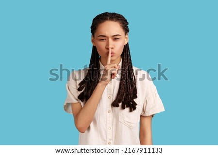Woman with black dreadlocks shushes with angry face, makes taboo gesture, tells to be quiet, demands not to spread rumors, scolds person being too loud. Indoor studio shot isolated on blue background.